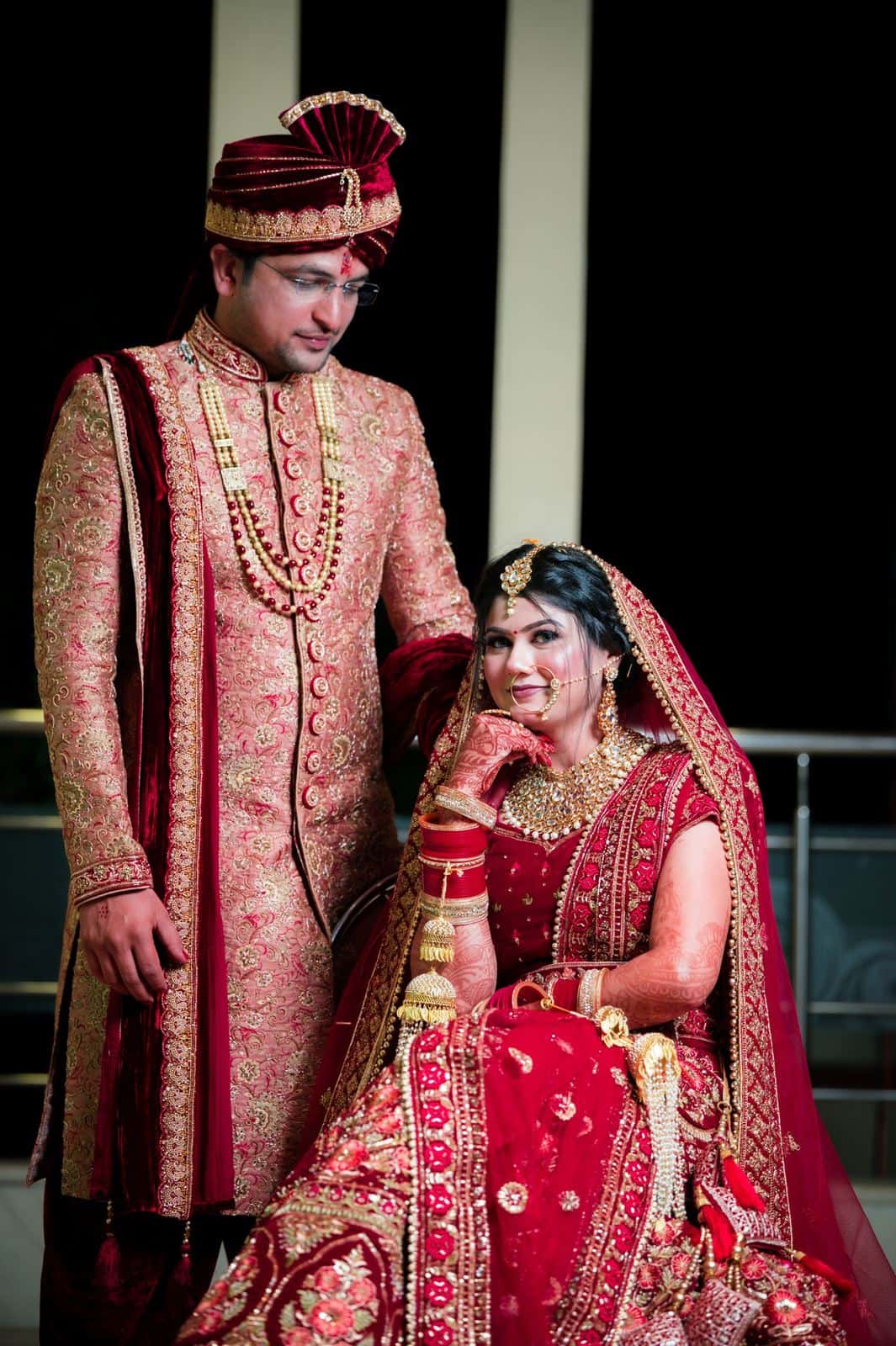 Traditional Indian bride in a regal red lehenga beside the groom in an embroidered cream sherwani and maroon safa